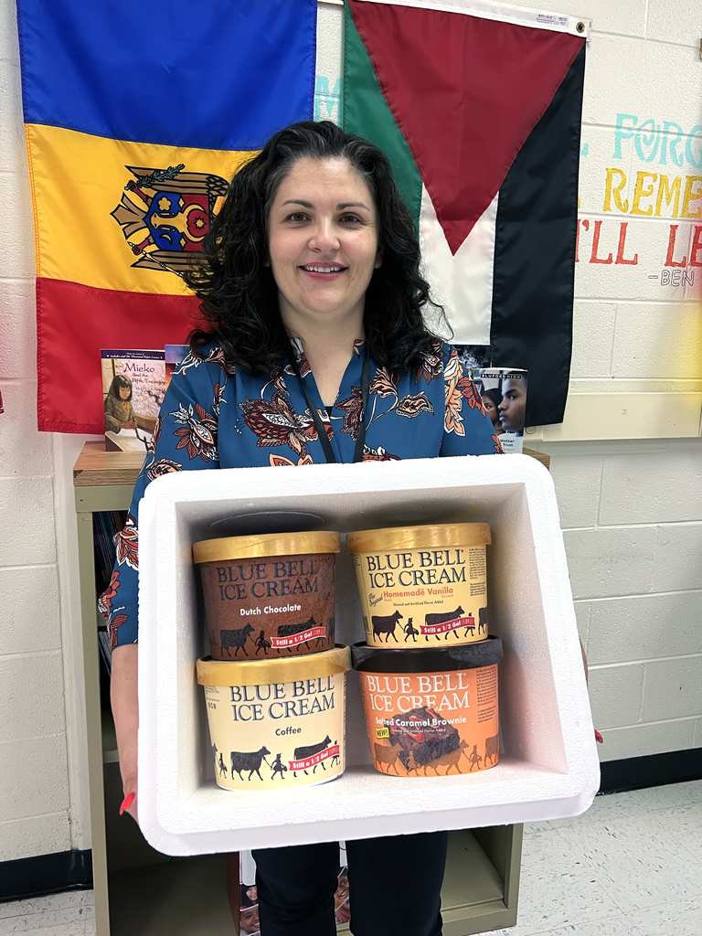 Ms. Fabiana Parker is standing in her classroom smiling and holding up a cooler with four half-gallon tubs of Blue Bell Ice Cream. The flavors are Dutch Chocolate, Coffee, Homemade Vanilla, and Salted Caramel Brownie.